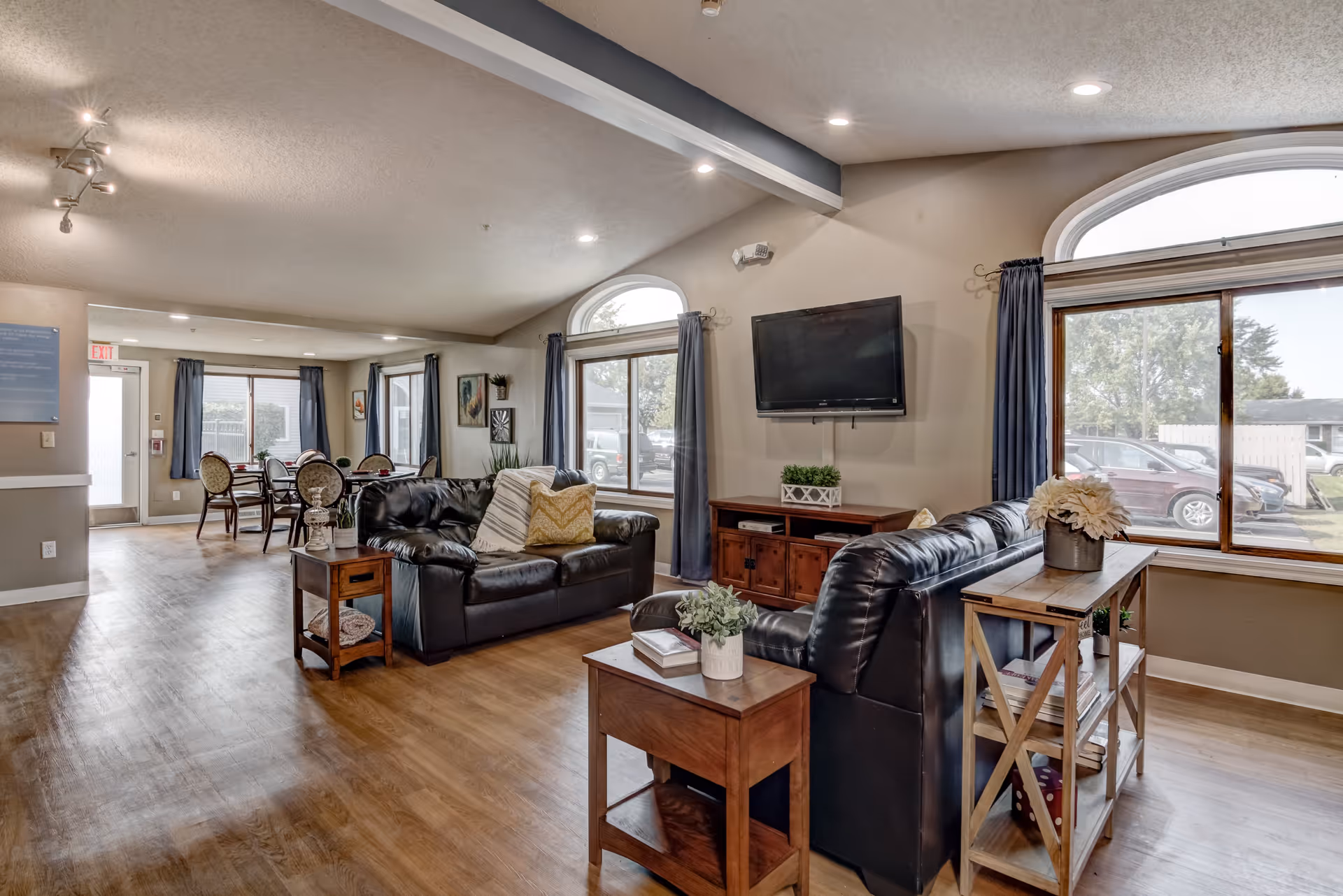 A spacious living room area in a senior living facility with two black leather sofas, wooden side tables with plants and books, a wall-mounted flat-screen TV, large windows with blue curtains, and a dining area with a table and chairs in the background.