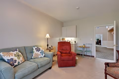 A cozy living room area with a light blue sofa adorned with floral pillows, a red recliner chair, a small wooden side table with a lamp, and a small round table with two chairs near a kitchenette. The walls are light-colored, and an open door leads to another room with carpeted flooring.