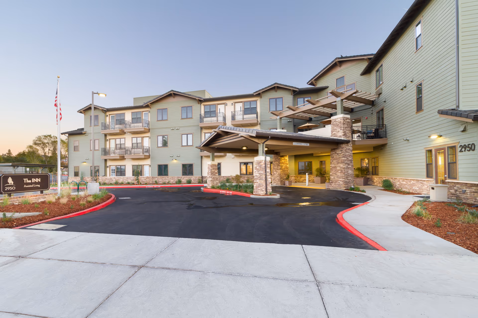 Exterior view of a multi-story assisted living facility named The Inn at 2950, with a covered entrance, balconies, and landscaped surroundings under a clear sky at dusk.