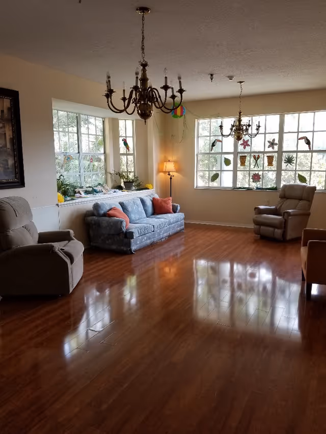 Sunny communal living room with sofas and recliners, hardwood floors, large decorated windows, and hanging chandeliers.