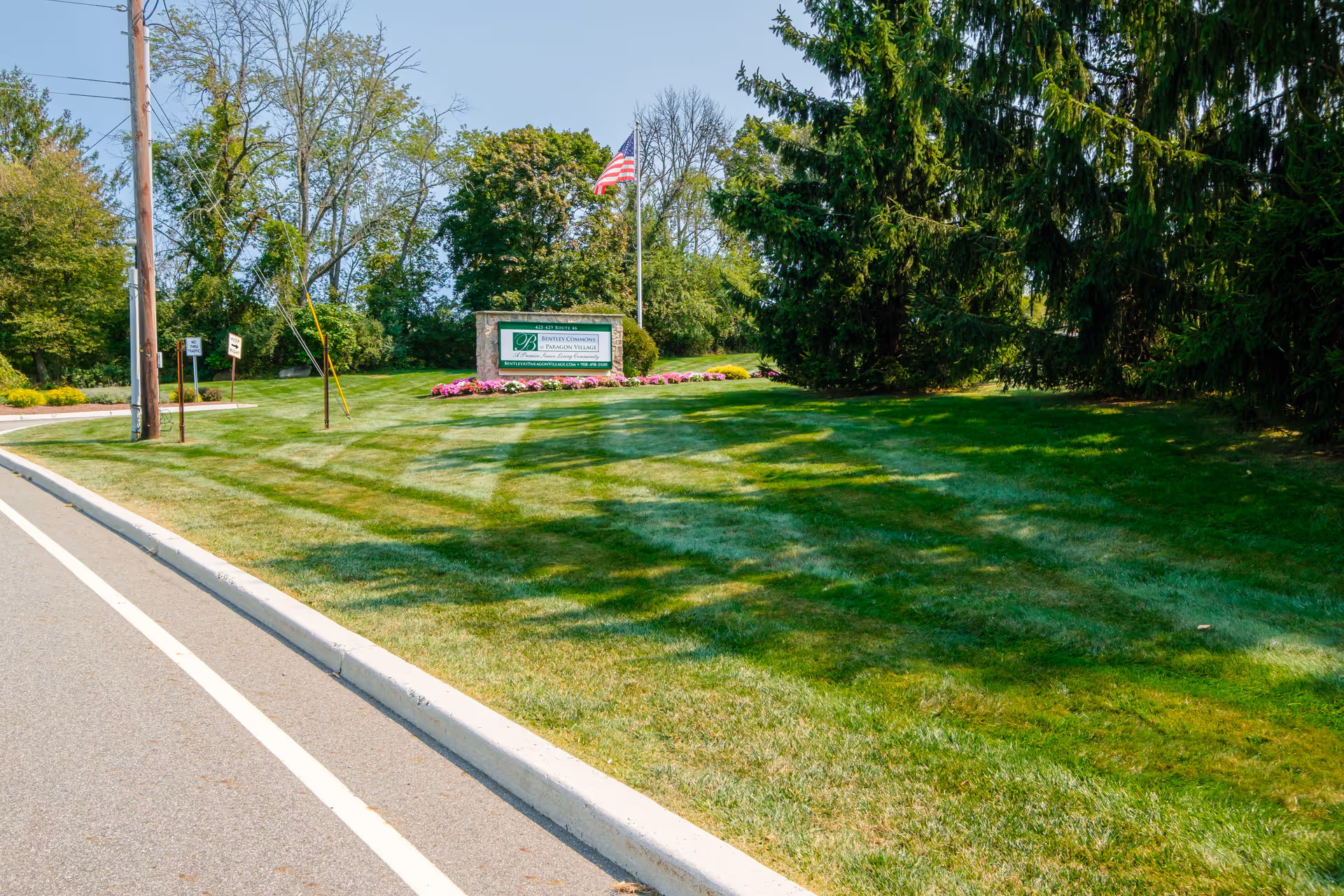 Entrance lawn with a stone sign for Bentley Commons at Paragon Village, an American flag, and manicured grass beside the road.
