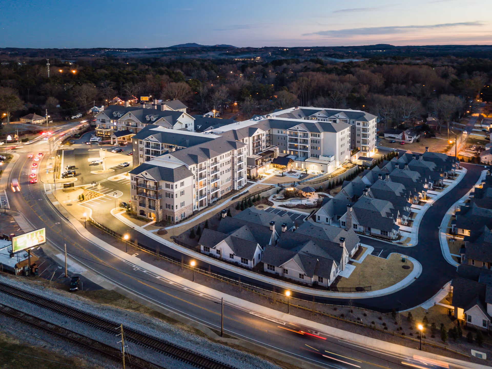Aerial view of the Holbrook Acworth senior living facility at dusk, showing multiple residential buildings with lit windows, surrounding roads with moving cars, and a landscaped courtyard area.