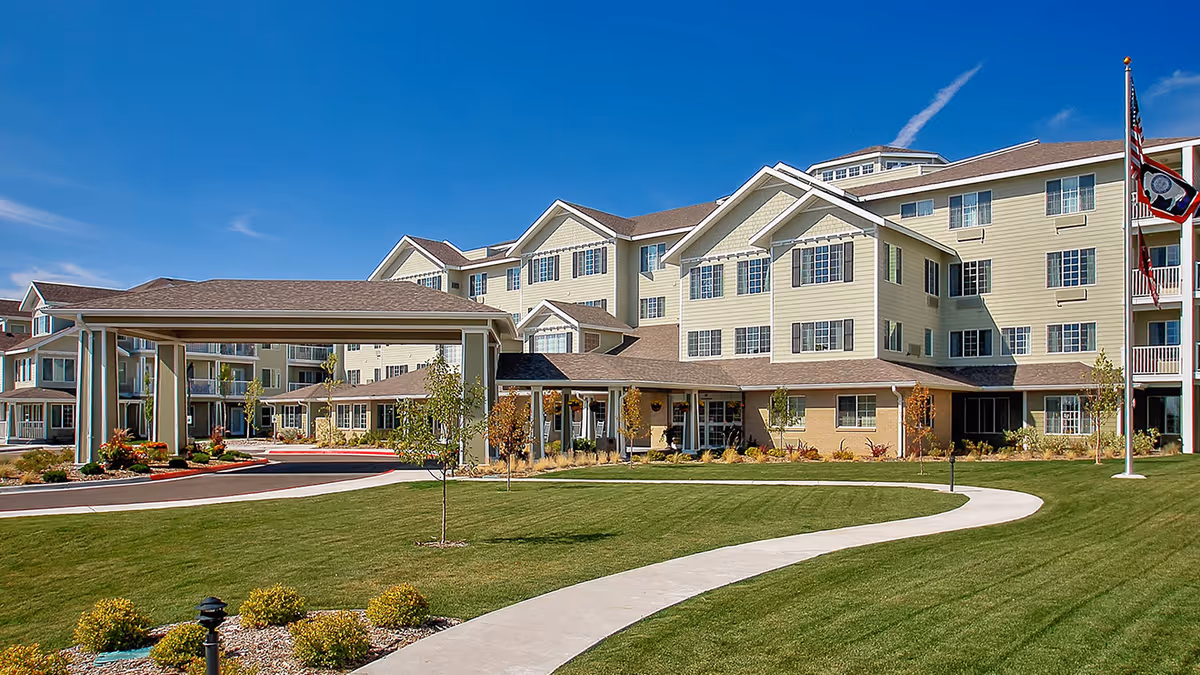 Exterior view of a large, multi-story senior living facility with beige siding and multiple windows under a clear blue sky. The building has a covered entrance with a driveway and a well-maintained lawn with a curved concrete walkway and small trees. Flags are visible on flagpoles to the right.