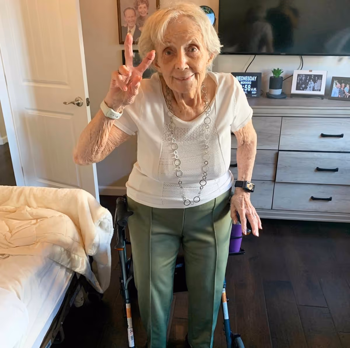 An elderly woman standing in a bedroom using a walker. She is wearing a white short-sleeve top, green pants, and a long necklace, and is making a peace sign with her right hand. Behind her is a dresser with framed photos, a small plant, and a digital clock. A bed with a beige blanket is visible on the left side of the image.