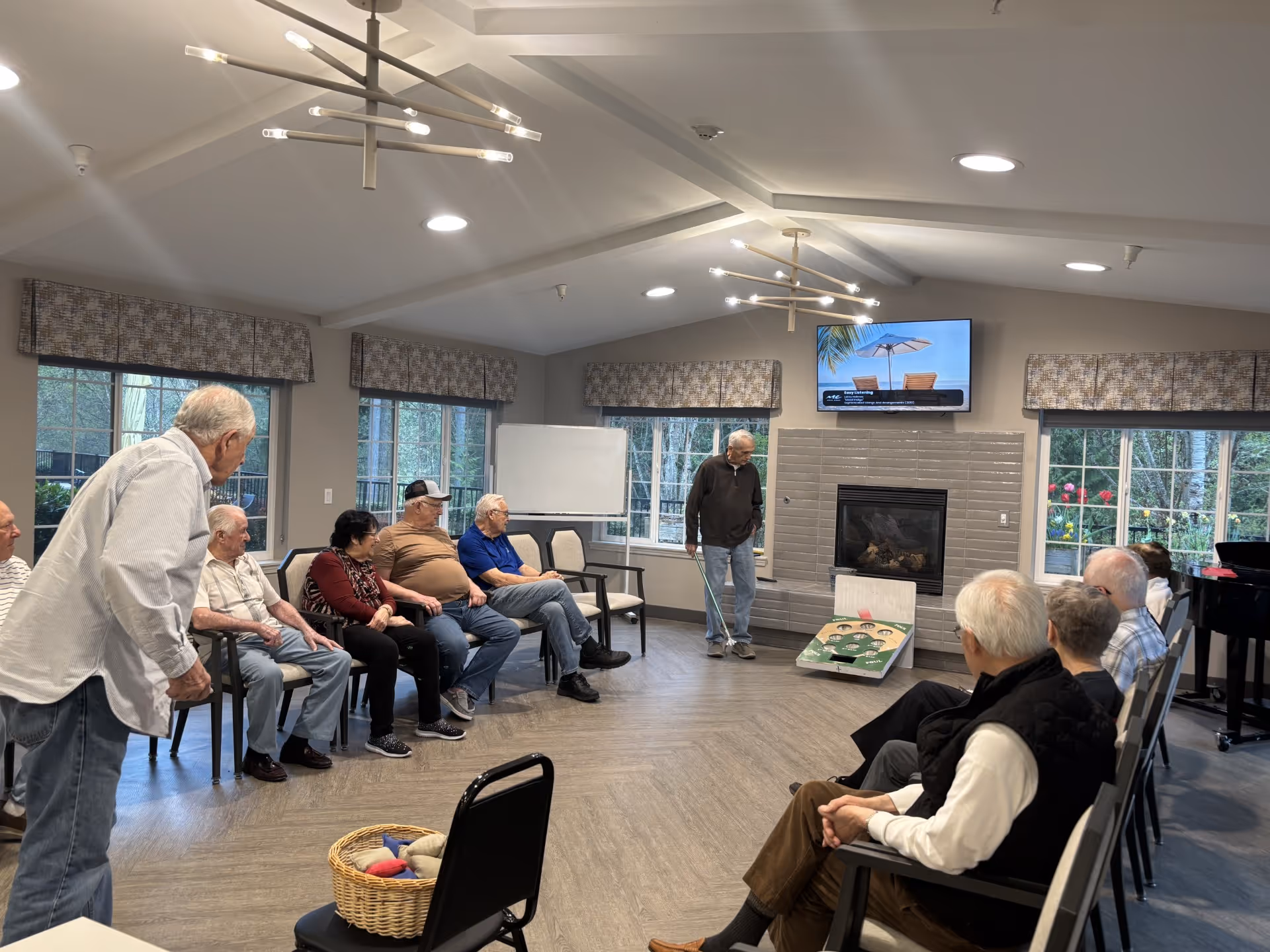 A group of elderly people sitting in a semi-circle in a well-lit room with large windows and modern ceiling lights. One man is standing and appears to be playing a bean bag toss game near a fireplace with a TV mounted above it. The room has a cozy and social atmosphere.