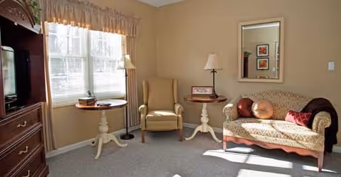 A cozy living room area with a beige armchair, a small round wooden table with a lamp, a loveseat with patterned upholstery and decorative pillows, a large window with sheer curtains allowing sunlight to fill the room, and a wooden entertainment cabinet.