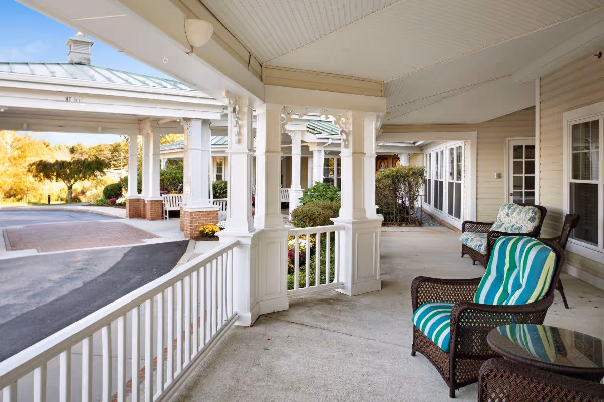 Covered front porch and entryway with wicker chairs and striped cushions overlooking the driveway at a senior living facility.