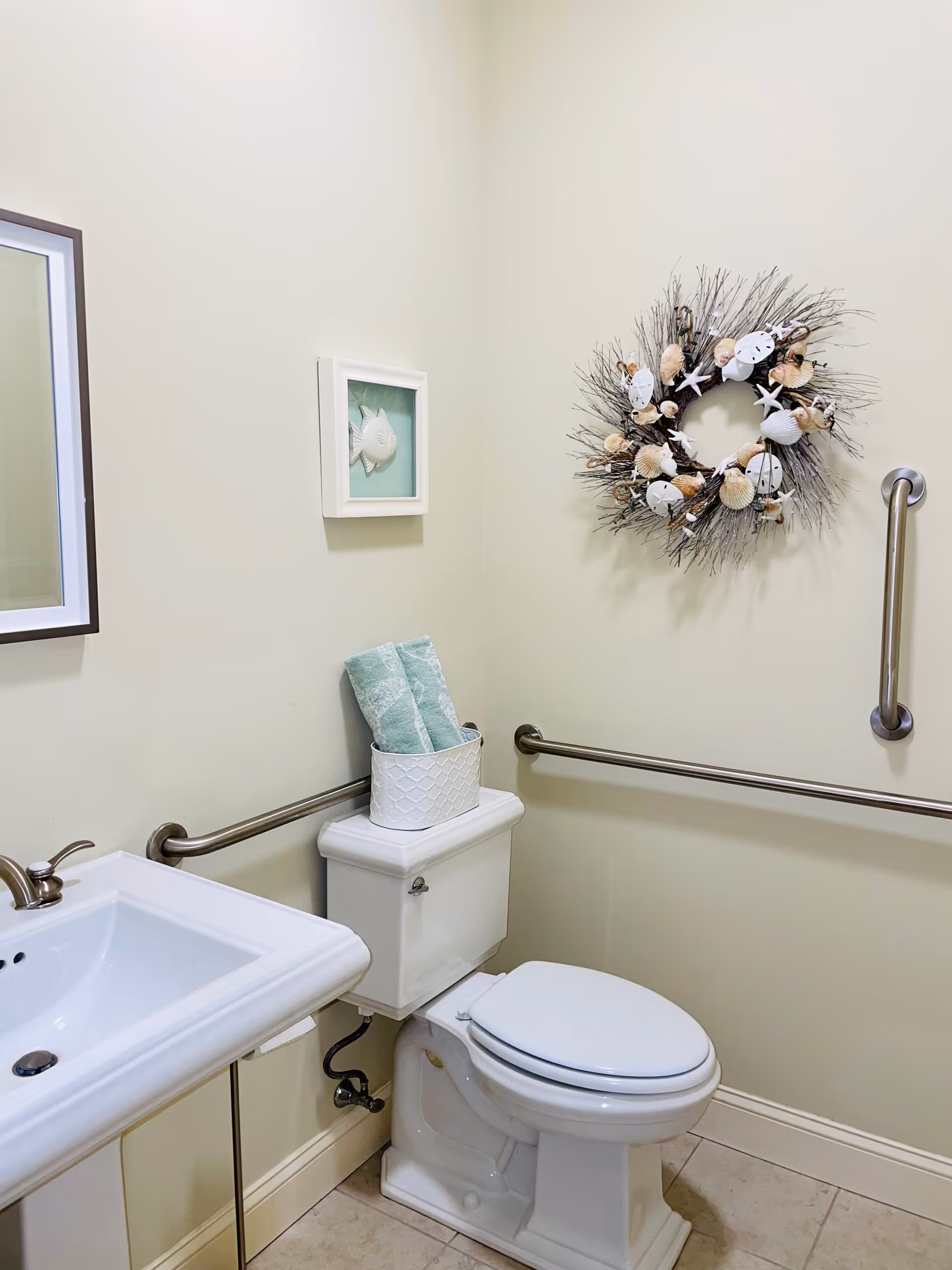 A clean and accessible bathroom with a white toilet and sink. The toilet tank has a white basket holding two folded green towels. There are stainless steel grab bars on the walls near the toilet for support. The walls are light-colored and decorated with a framed fish artwork and a wreath made of twigs, seashells, and starfish.