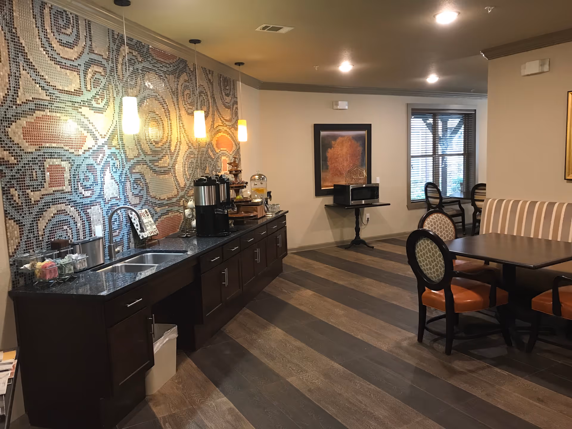 Interior view of a communal dining and kitchen area with a long counter featuring a sink, coffee dispensers, and various condiments. The wall behind the counter has a colorful mosaic tile design. There are pendant lights hanging above the counter. In the background, there is a table with chairs and a striped bench seat, a microwave on a small table, and a window with blinds letting in natural light.