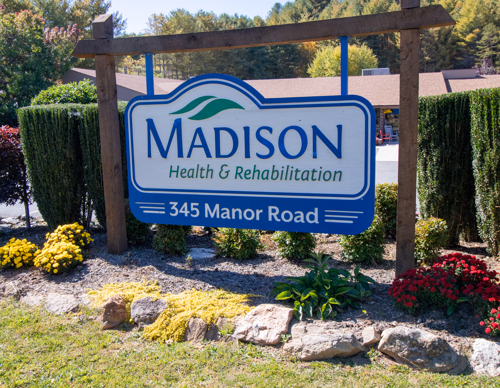 A blue and white wooden sign reading "Madison Health & Rehabilitation 345 Manor Road" in a landscaped entrance with flowers and trimmed hedges.