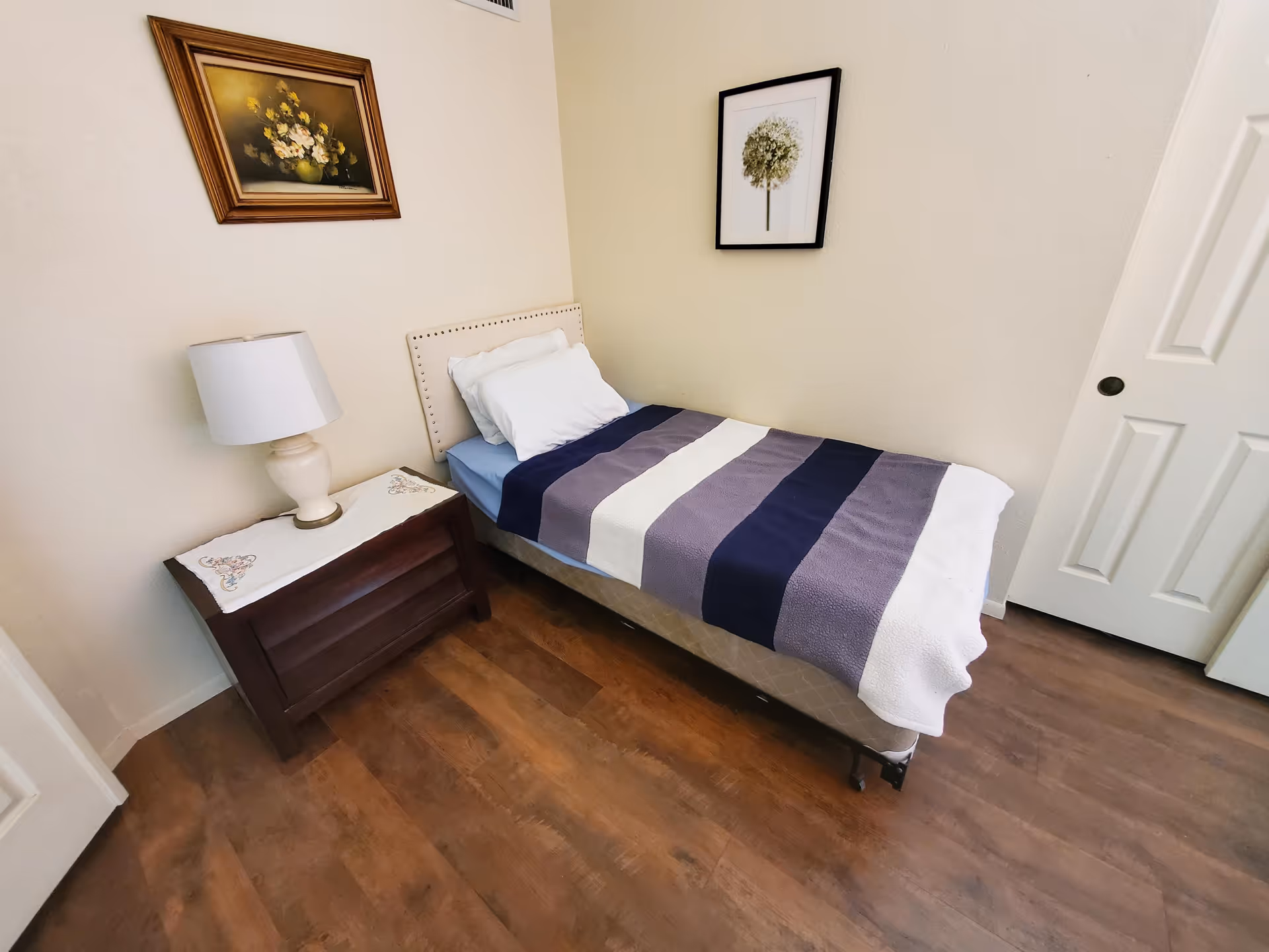 A small bedroom with a single bed covered with a striped blanket in shades of blue, gray, and white. There is a white pillow and a blue fitted sheet on the bed. Next to the bed is a wooden nightstand with a white table lamp on top. The walls are light-colored and decorated with two framed pictures, one of flowers and the other of a tree. The floor is wooden, and there are two white doors visible in the room.