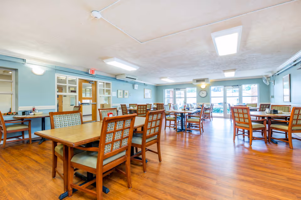 A bright dining room with multiple wooden tables and chairs arranged neatly on a wooden floor. The walls are painted light blue and decorated with framed pictures. Large windows and glass doors let in natural light, and there is a clock on the wall near the doors.