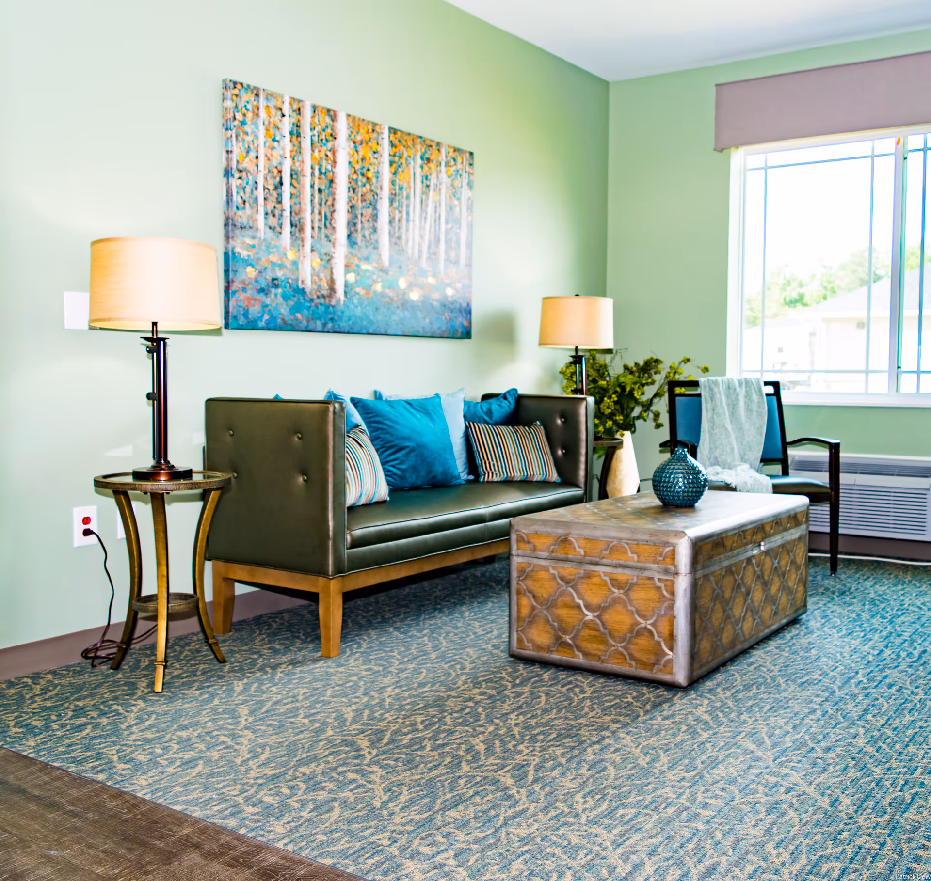 A cozy living room with a green leather sofa adorned with blue and striped pillows, two side tables each with a lamp, a decorative wooden coffee table, a chair with a light blue throw blanket, a large window with a view outside, and a colorful painting of birch trees on a light green wall.