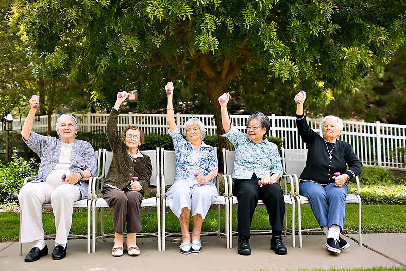 Five elderly women sitting outdoors on chairs in a row, each holding a small purple dumbbell and raising one arm in the air, with green trees and a white fence in the background.