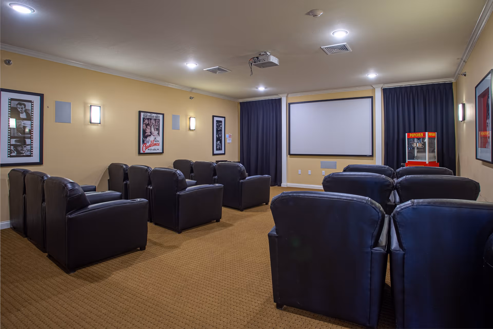 A small movie theater room with multiple rows of black leather recliner chairs facing a large white projection screen. The walls are painted beige and decorated with framed classic movie posters. A popcorn machine is visible in the back corner of the room. Ceiling lights and a projector are mounted on the ceiling.