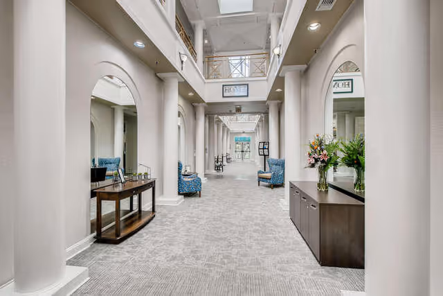 A bright and spacious hallway in a senior living facility with high ceilings and skylights. The hallway features large mirrors on both sides, decorative columns, and comfortable blue armchairs placed along the walls. There are tables with flower arrangements and a carpeted floor, creating a welcoming and elegant atmosphere.