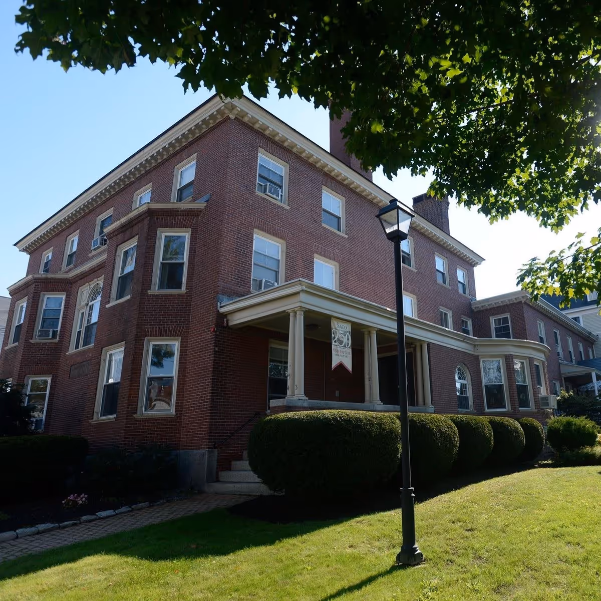 Exterior view of a large, three-story brick building with multiple windows and a covered entrance supported by columns. The building is surrounded by neatly trimmed bushes and a green lawn, with a black lamppost in front and tree branches partially shading the scene.