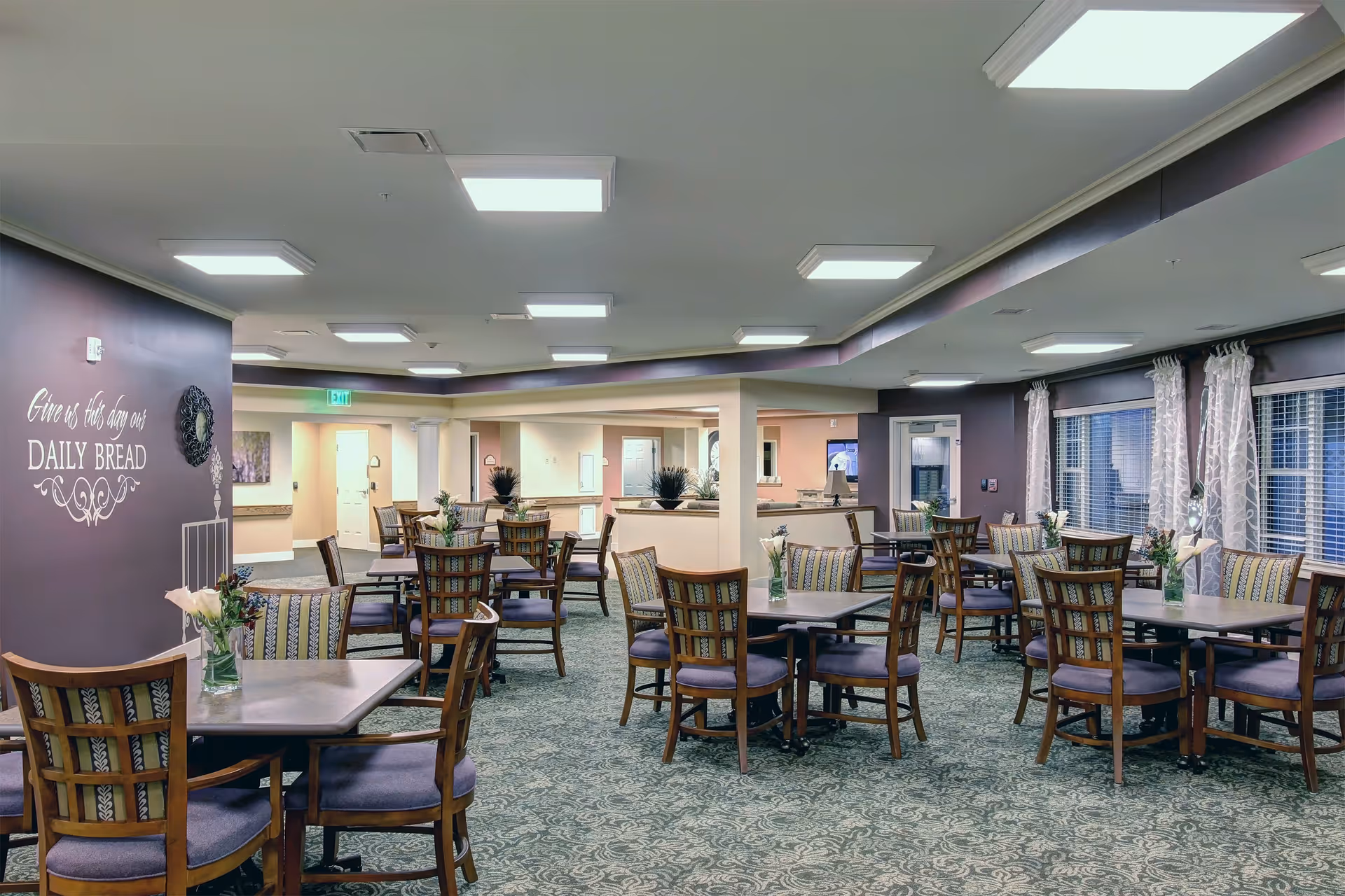 A spacious dining room with multiple tables and wooden chairs arranged neatly. Each table has a small vase with flowers. The room has patterned carpet flooring, large windows with sheer curtains, and soft overhead lighting. On one wall, there is a decorative sign that reads 'Give us this day our DAILY BREAD'.