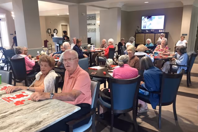 A group of elderly residents seated at multiple tables in a bright communal dining/activity room, playing games and socializing.