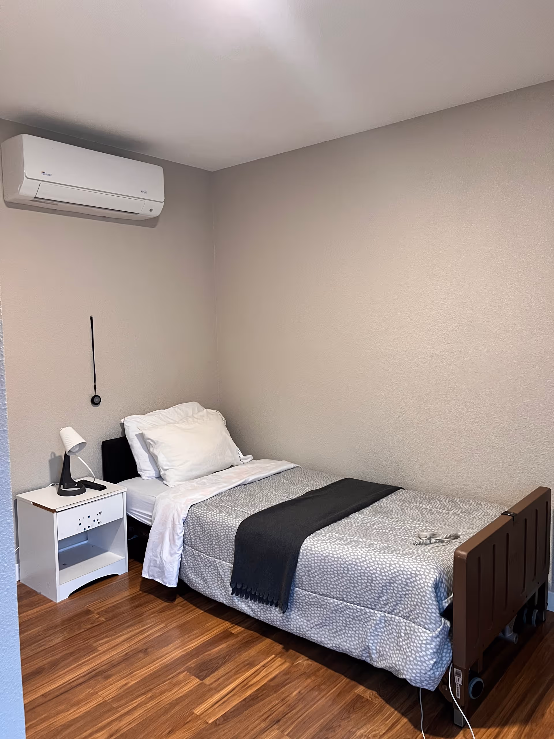 A small bedroom with a single bed, nightstand and lamp, hardwood floor, and a wall-mounted air conditioner.