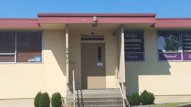 Exterior front view of a single-story building with beige walls and a red roof trim. The entrance has a small staircase with railings leading to a door under a covered porch area. Signs near the door read 'Home Instead Senior Care' and 'HCAC Training'. There are windows on either side of the entrance, and small bushes are planted along the front of the building.