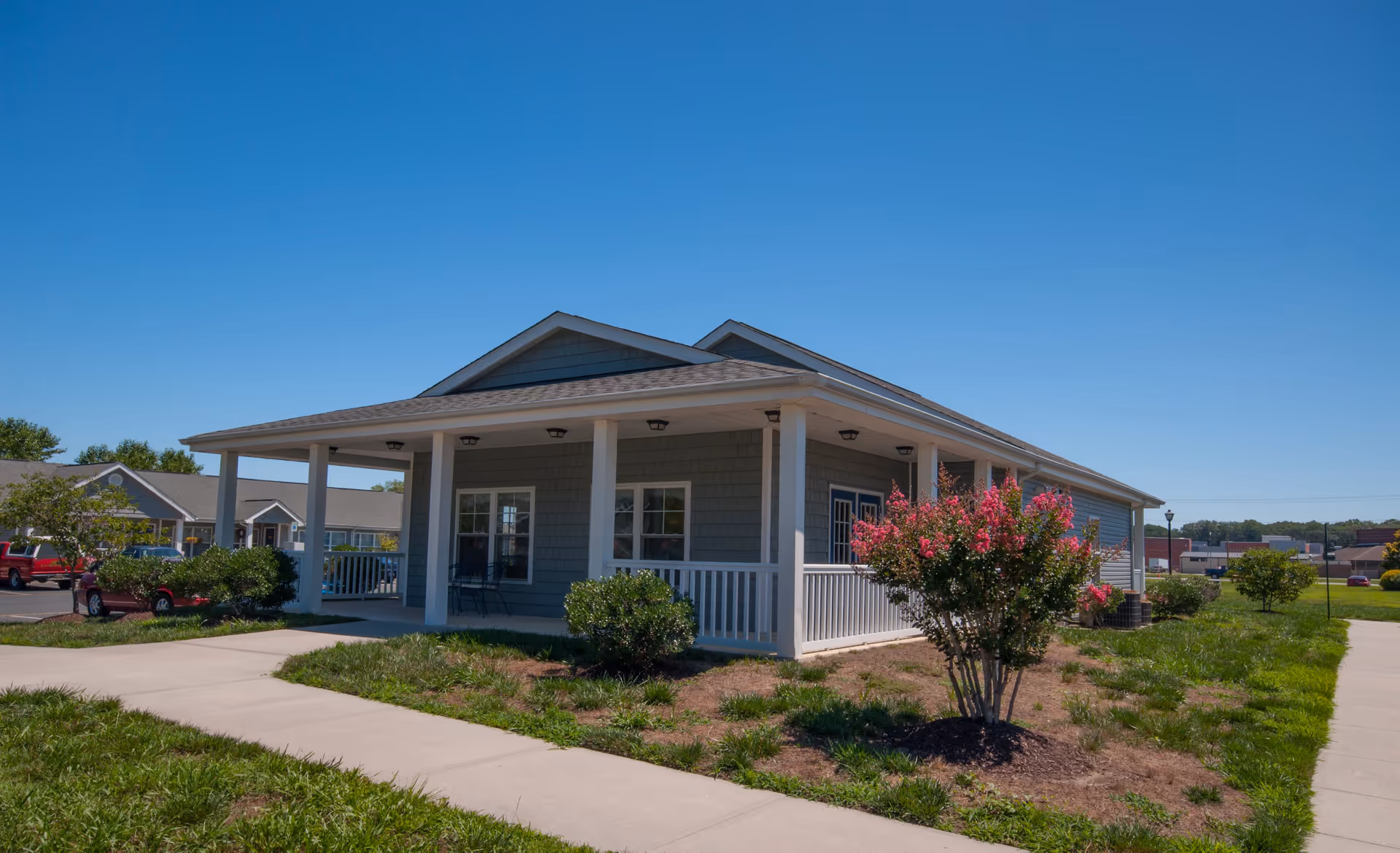 Single-story senior living building with a covered porch, white columns, and landscaped shrubs under a clear blue sky.
