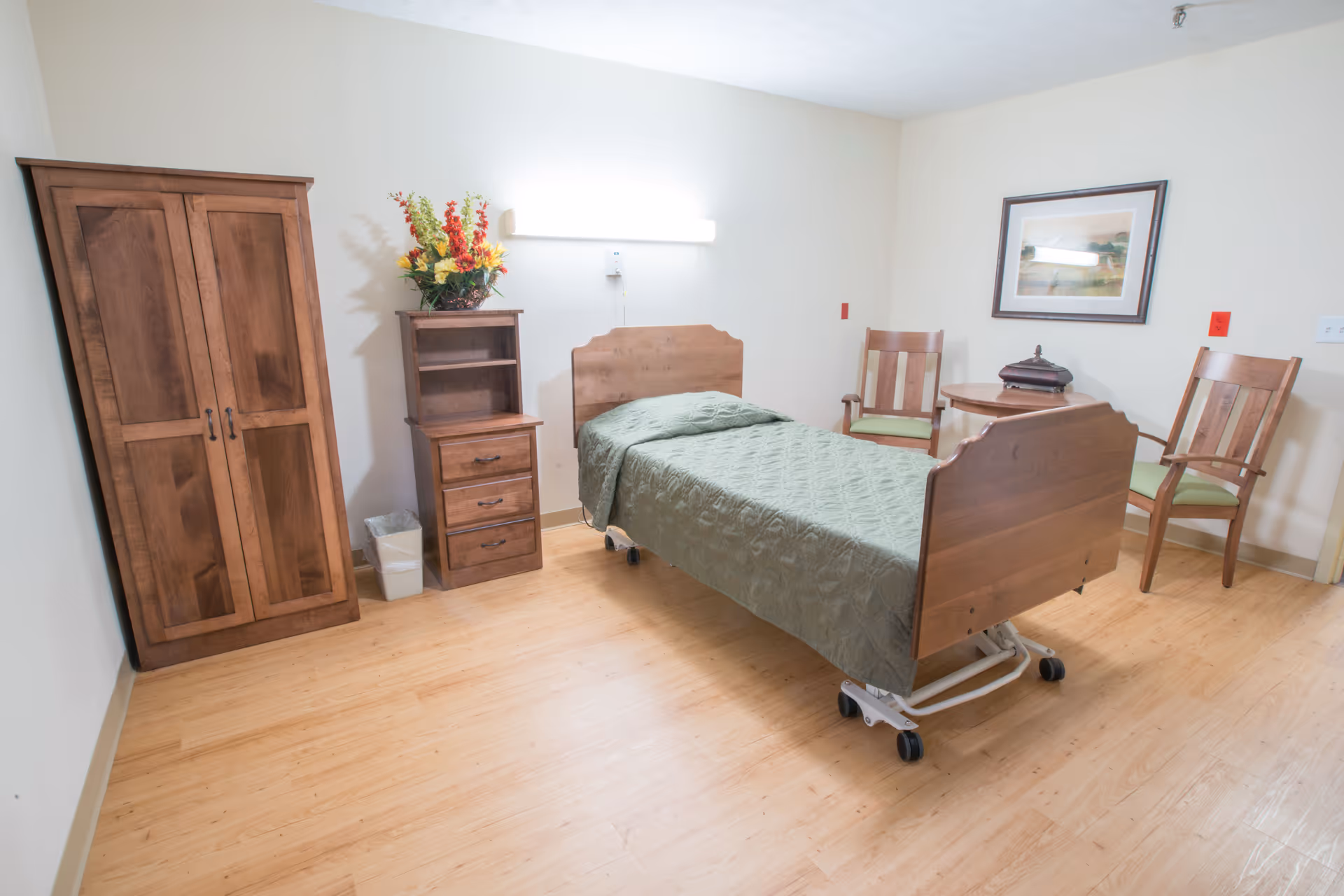 A simple, clean bedroom in a care facility featuring a single wooden bed with a green quilt, a wooden wardrobe, a small wooden dresser with a flower arrangement on top, two wooden chairs with green cushions, a small round table, and a framed picture on the wall.