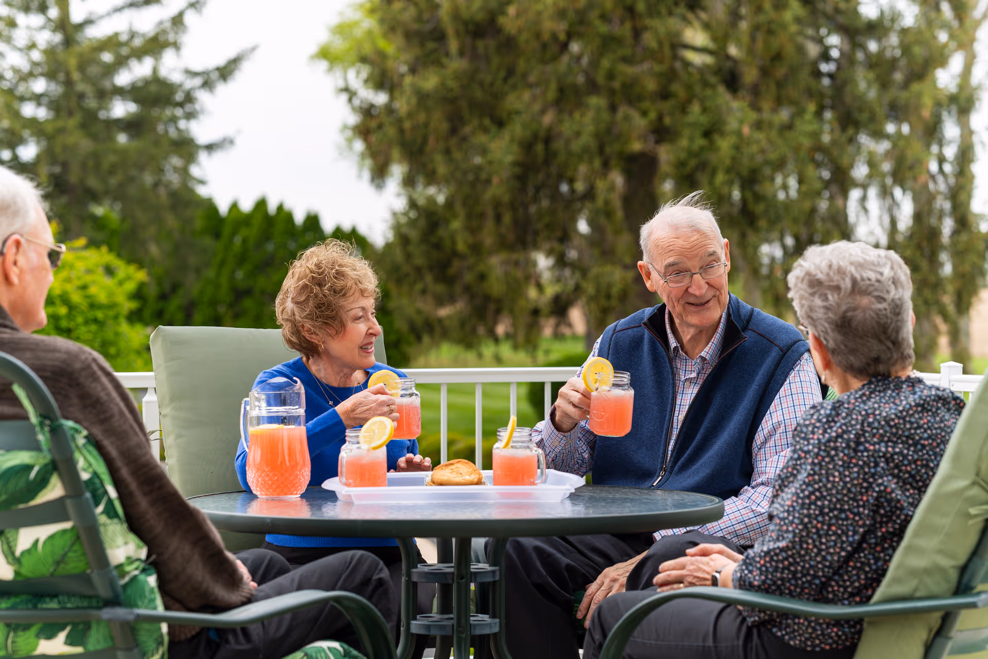 Four elderly adults sit around a patio table outdoors, enjoying pink lemonade with lemon slices and pastries while chatting.
