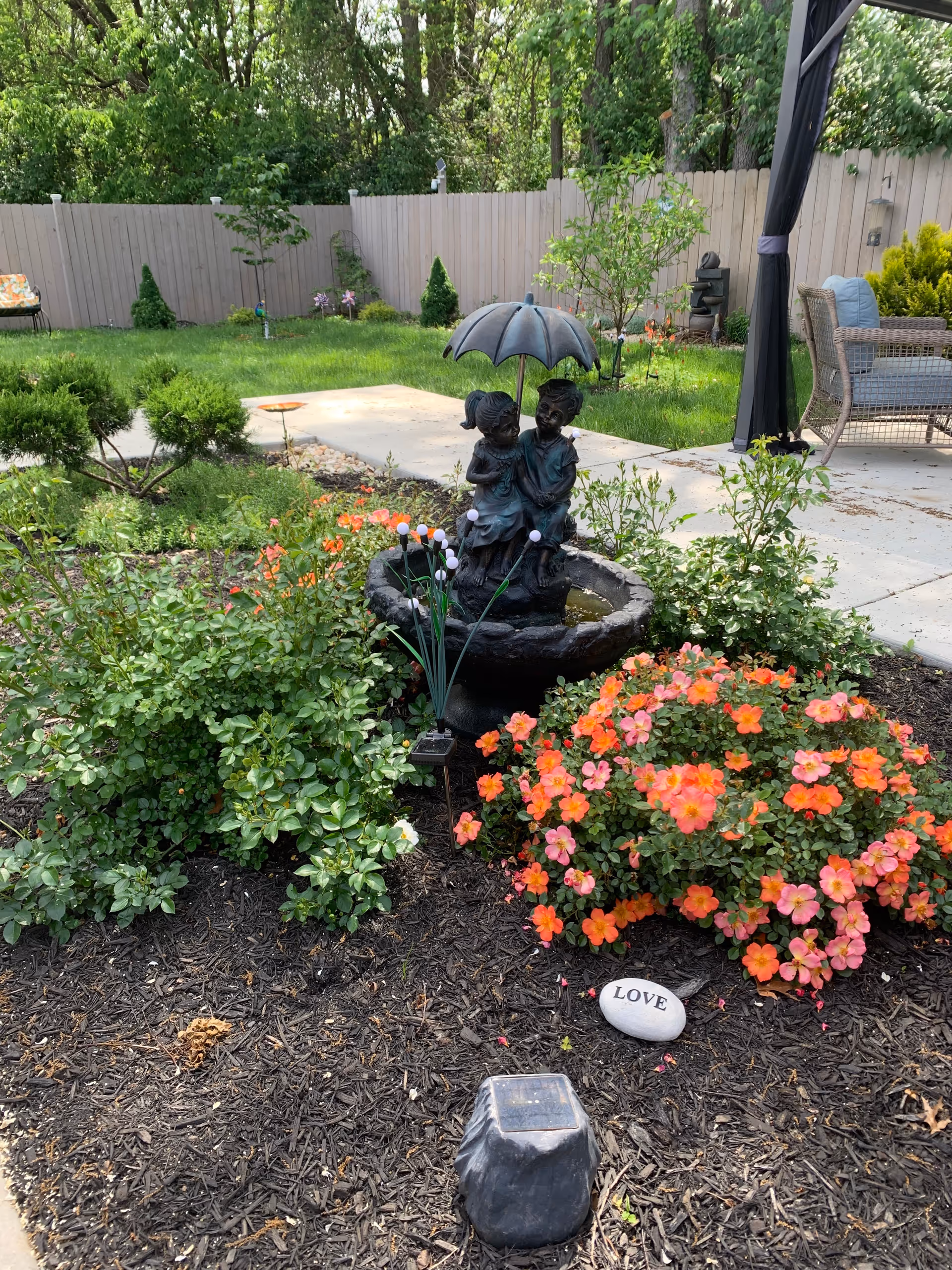 A garden area with a statue of two children sitting under an umbrella in the center of a small water fountain. Surrounding the fountain are green bushes and vibrant orange flowers. A white stone with the word 'LOVE' is placed on the mulch in front of the flowers. In the background, there is a wooden fence, a concrete patio with outdoor furniture, and trees providing greenery.