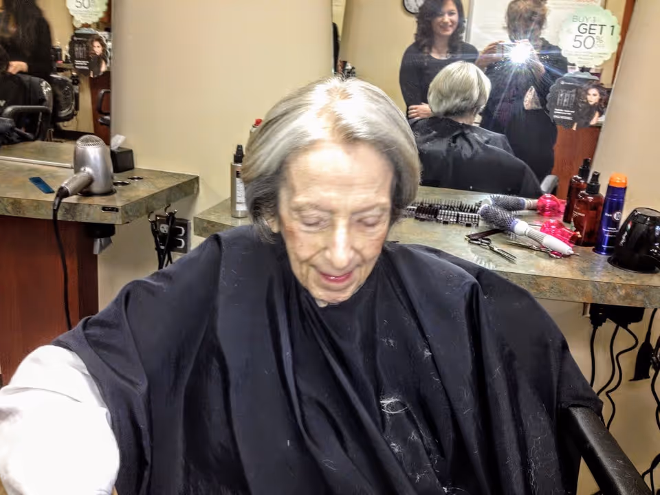An elderly woman with gray hair is seated in a salon chair wearing a black cape, with a hairdresser standing behind her. The background shows a mirror reflecting the woman and the hairdresser, along with various hair styling tools and products on the counter.