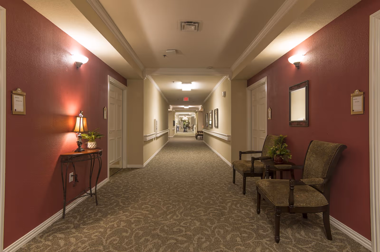 A long, well-lit hallway in an assisted living facility with beige and maroon walls. The hallway is carpeted with a patterned design and features several doors on both sides. There are two upholstered chairs with a small table and plant on the right side, and a small table with a lamp and plant on the left side. Wall sconces provide additional lighting, and framed pictures and a mirror decorate the walls.