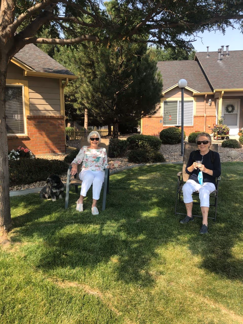 Two elderly women sitting on chairs on a grassy lawn outside a residential building, both wearing sunglasses and holding drinks. There is a small black dog sitting on the grass near one of the women. The background shows trees, bushes, and parts of the building with brick and siding exterior.