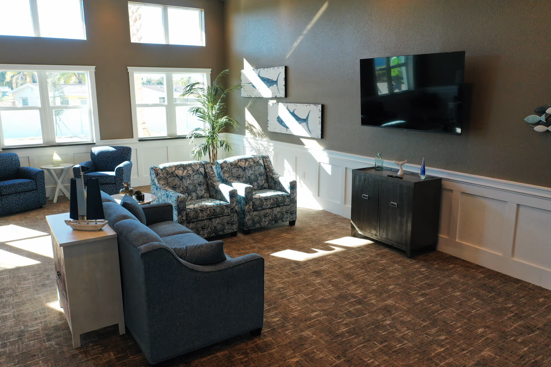 Bright senior living lounge with blue sofas, chairs, a TV, and windows.
