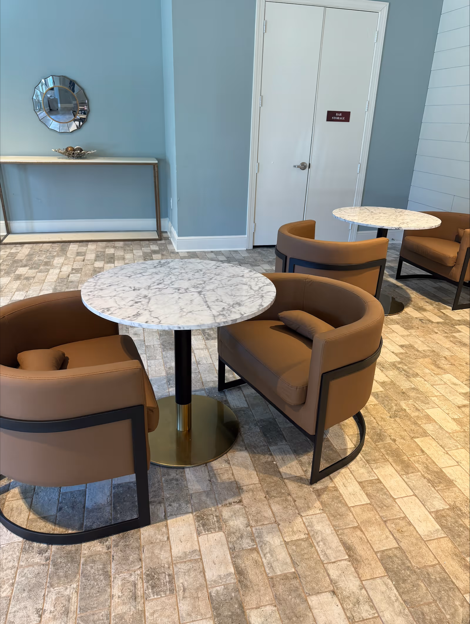 Interior seating area with two round marble-top tables, each surrounded by two brown cushioned armchairs with black frames. The floor is covered with light-colored brick-patterned tiles. In the background, there is a light blue wall with a decorative round mirror above a narrow console table. A white double door with a sign labeled 'BAR STORAGE' is also visible.