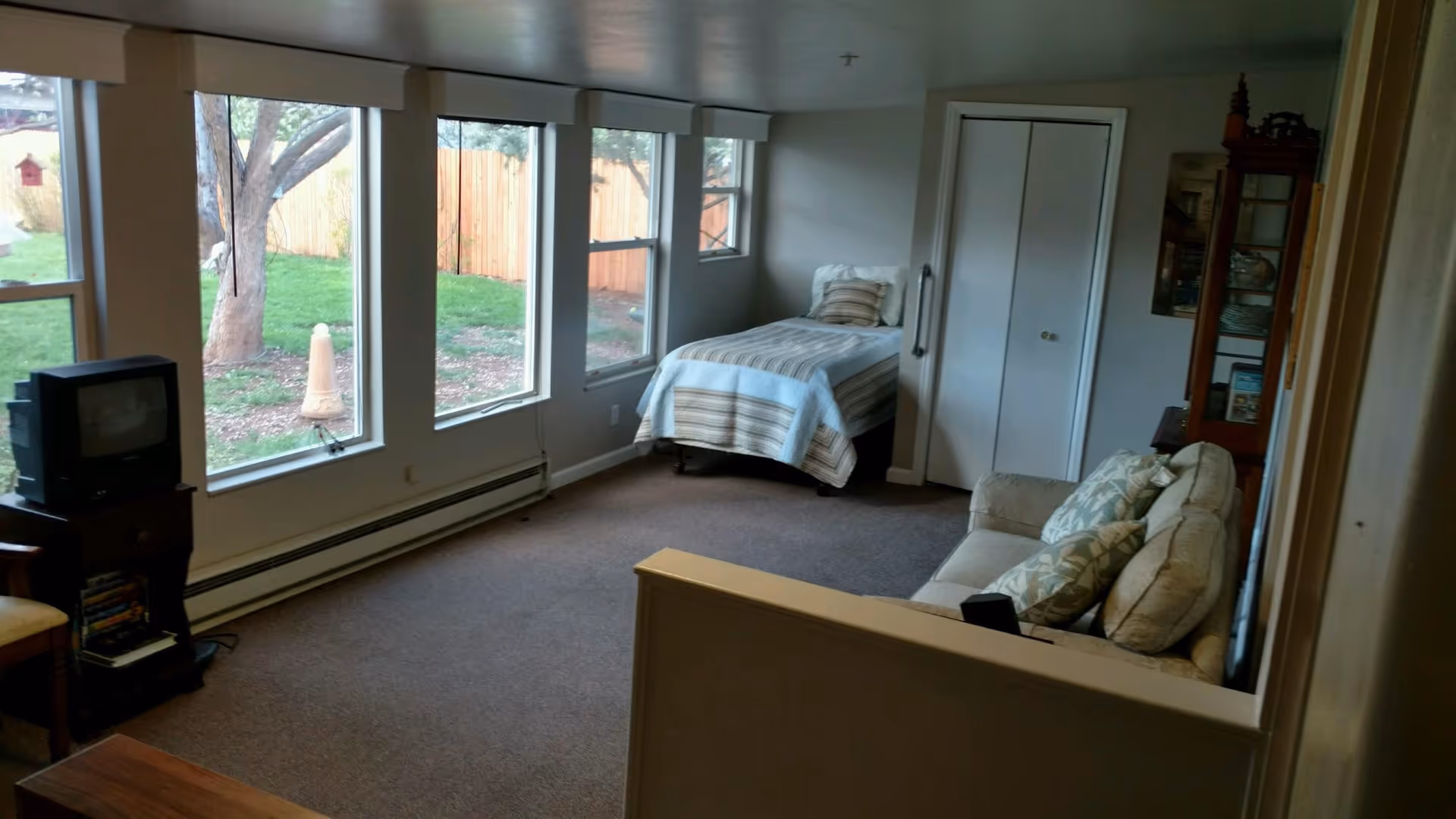 A room with a single bed covered with a striped blanket near a window, a beige couch with patterned pillows, a wooden cabinet with glass doors, and a small TV on a stand. Large windows show a green outdoor area with trees and a wooden fence.