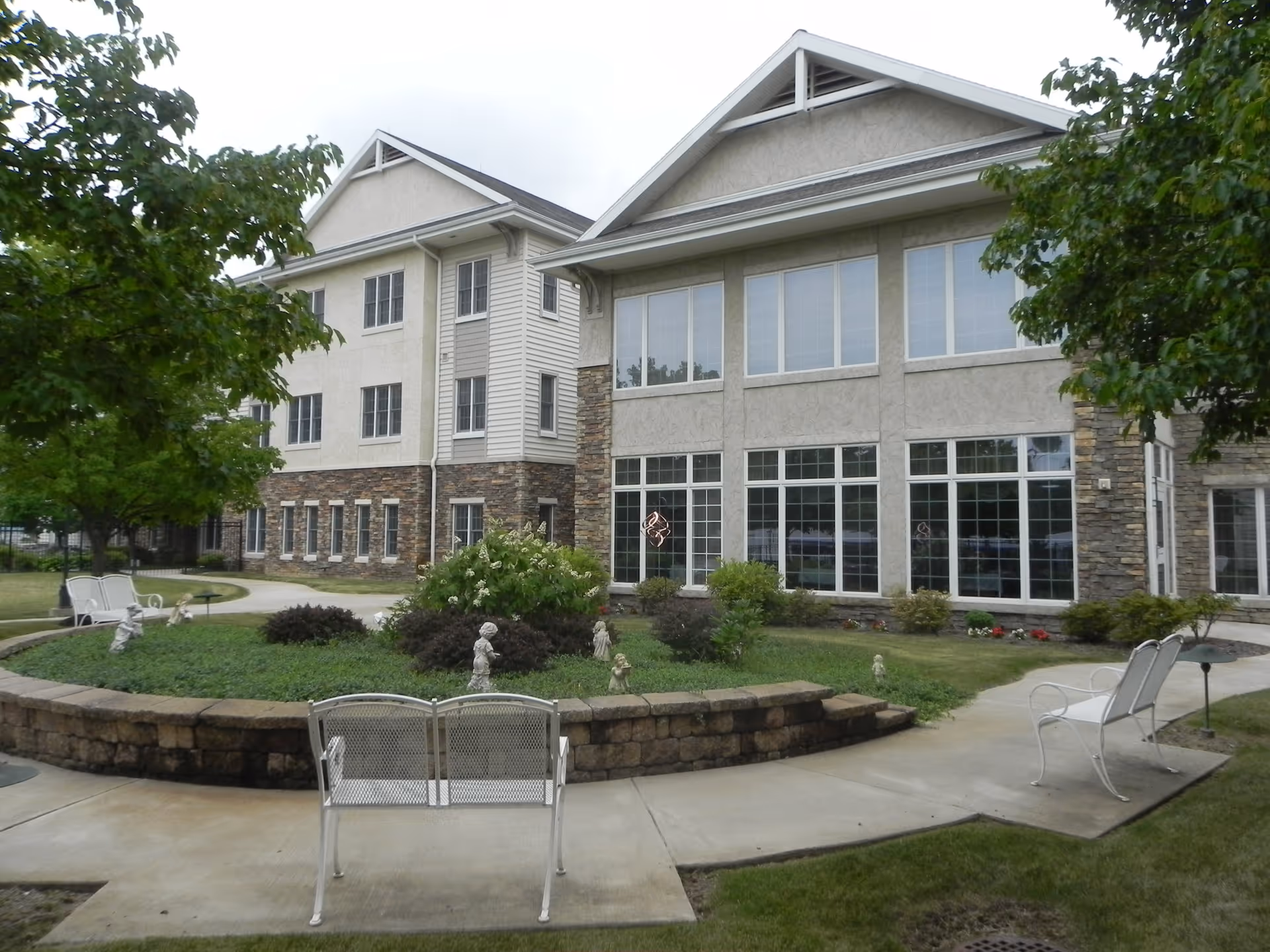 Outdoor view of a multi-story nursing home building with large windows, surrounded by a landscaped garden area with trees, shrubs, small statues, and white metal benches along a curved concrete pathway.