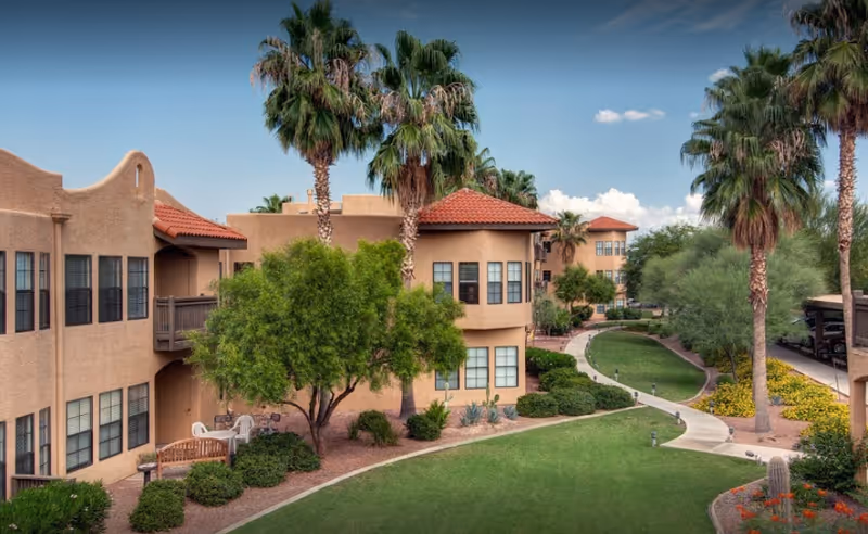 Exterior view of Amber Lights senior living campus with tan buildings, palm trees, and green lawns.