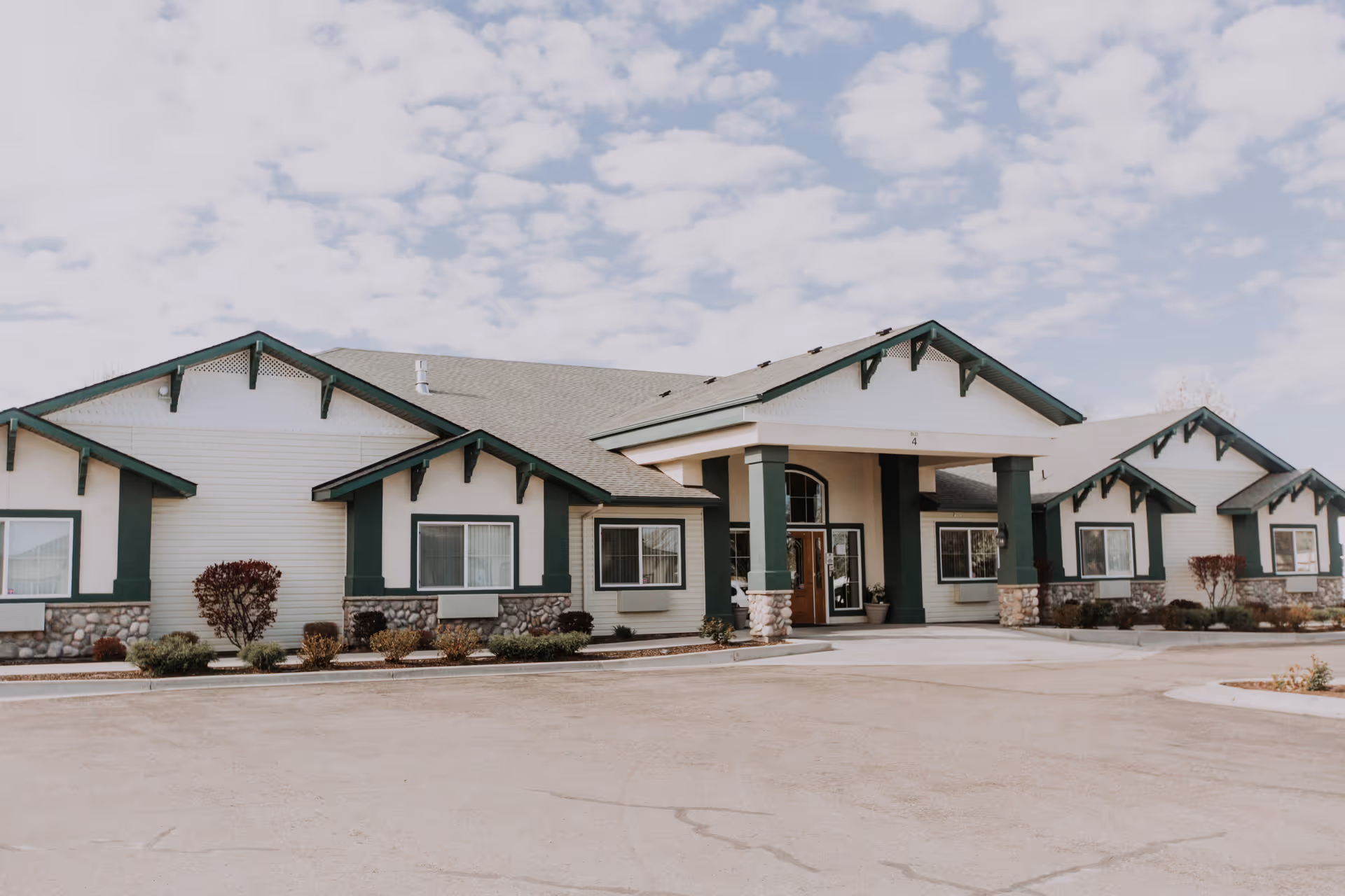 Exterior front view of a single-story assisted living facility building with green trim and stone accents under a partly cloudy sky. The entrance has a covered porch supported by columns, and there are shrubs and small plants along the front of the building.