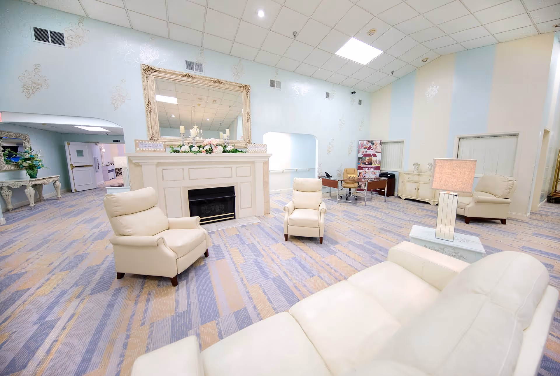 A spacious, well-lit common area with cream-colored leather armchairs and sofas arranged around a white fireplace with a large ornate mirror above it. The room has pastel-colored walls with subtle decorative patterns, a patterned carpet in shades of blue and beige, and a desk with a chair and promotional banner in the background.