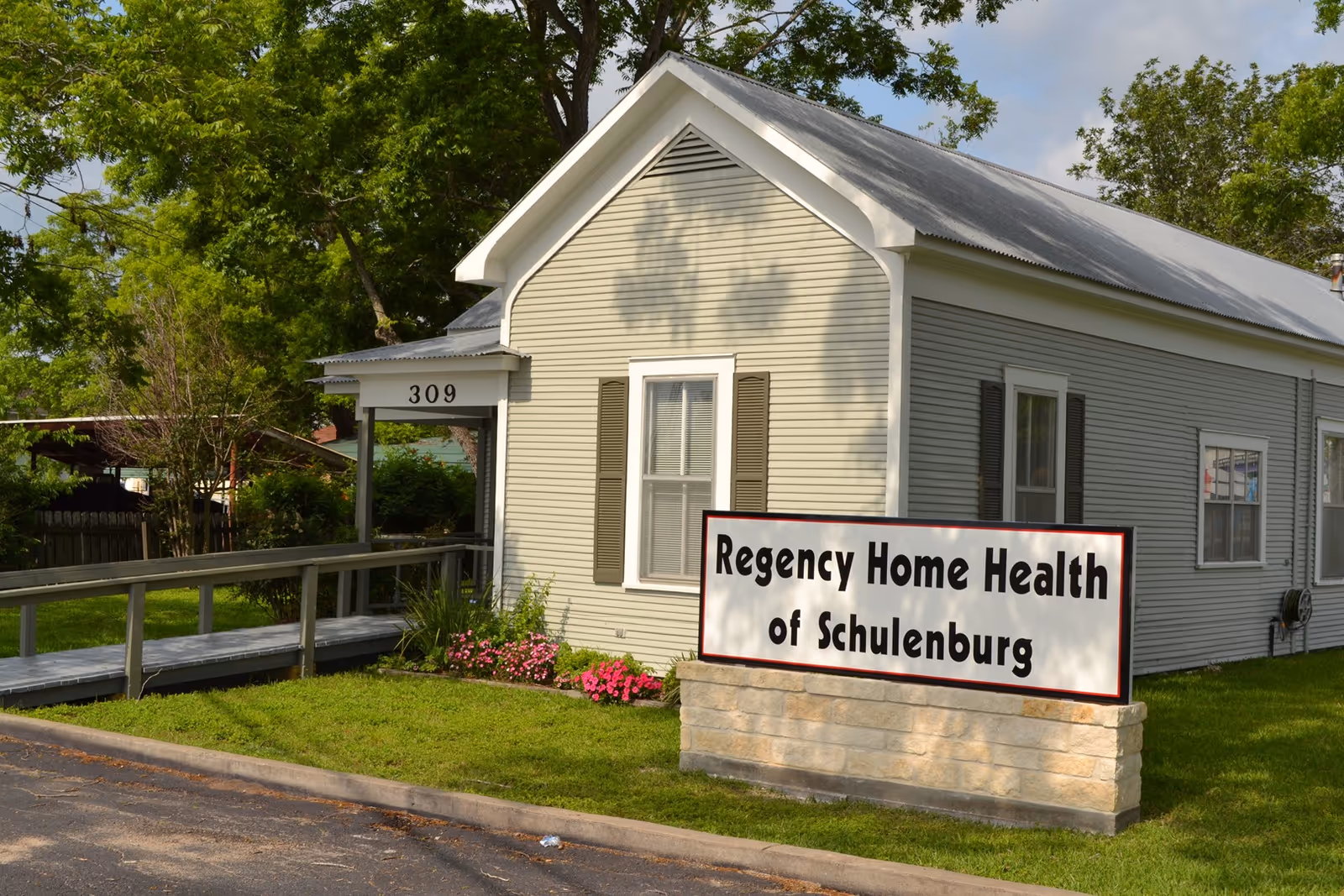 Exterior view of a single-story light gray building with white trim and a metal roof. There is a ramp leading to the entrance, which has the number 309 above it. In front of the building, there is a sign that reads 'Regency Home Health of Schulenburg' surrounded by green grass and some pink flowers.