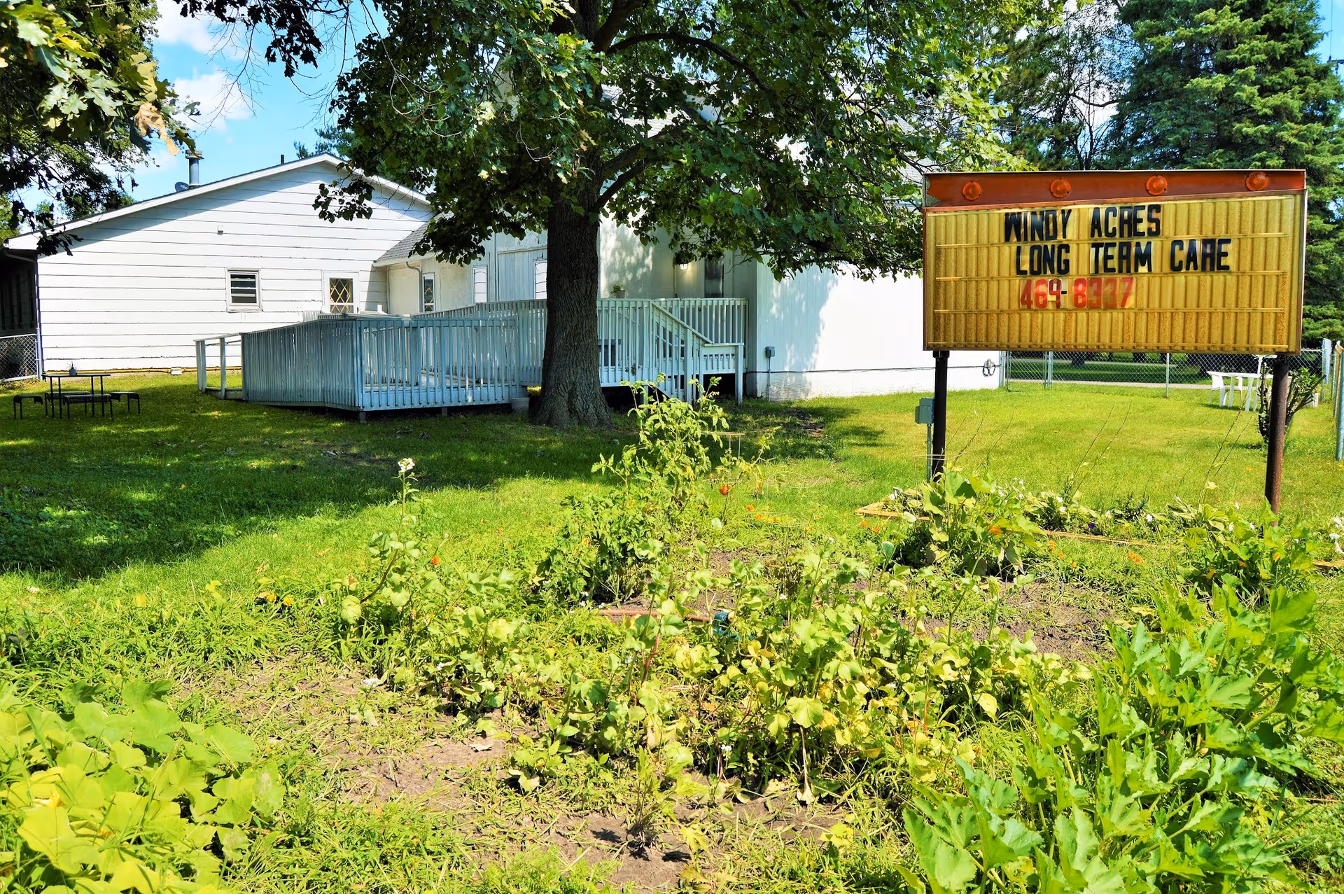 Outdoor view of Windy Acres Long Term Care Inc. showing a grassy yard with plants and a large tree. There is a white building with a wheelchair ramp and a yellow sign displaying the facility's name and phone number.