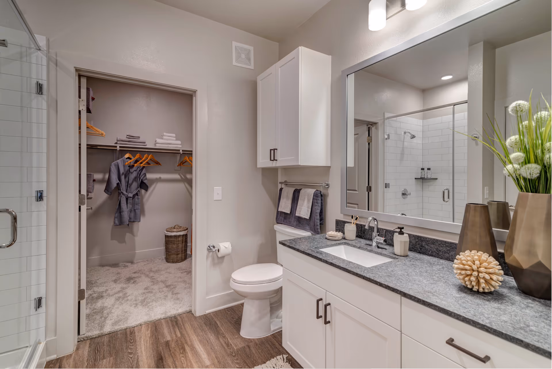 A modern bathroom featuring a white toilet, a gray countertop with a sink, and a large mirror above. There are decorative items including two bronze vases with white flowers and a spiky beige ornament on the countertop. A towel rack with folded towels is mounted above the toilet. To the left, there is a glass-enclosed shower with white subway tiles. An open doorway leads to a carpeted walk-in closet with hanging robes, towels, and a wicker laundry basket.