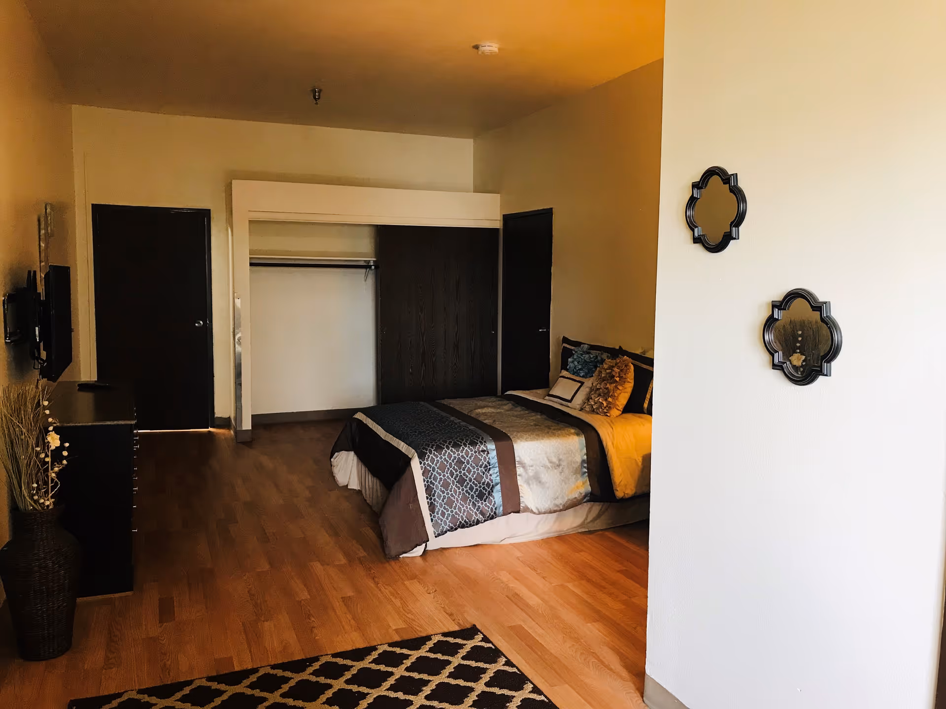 Studio-style bedroom with a made bed, open closet, dresser with TV, wood floors, and decorative wall mirrors.