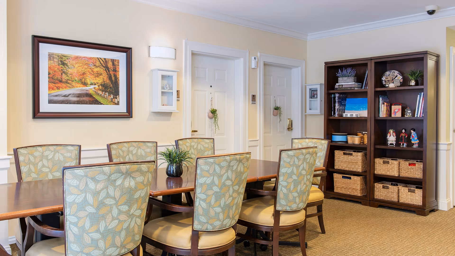 A well-lit dining room with a wooden table surrounded by six upholstered chairs with leaf-patterned backs. On the table is a small potted plant. The walls are light-colored with white trim, featuring a framed picture of a scenic autumn road and two white wall-mounted cabinets. In the background, there are two white doors with small hanging plants and a dark wooden bookshelf filled with books, baskets, and decorative items.