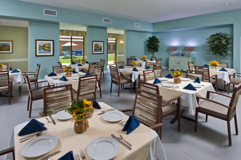 Bright dining room with multiple tables set with white tablecloths, navy napkins, plates, silverware and floral centerpieces, with windows looking out to a courtyard.