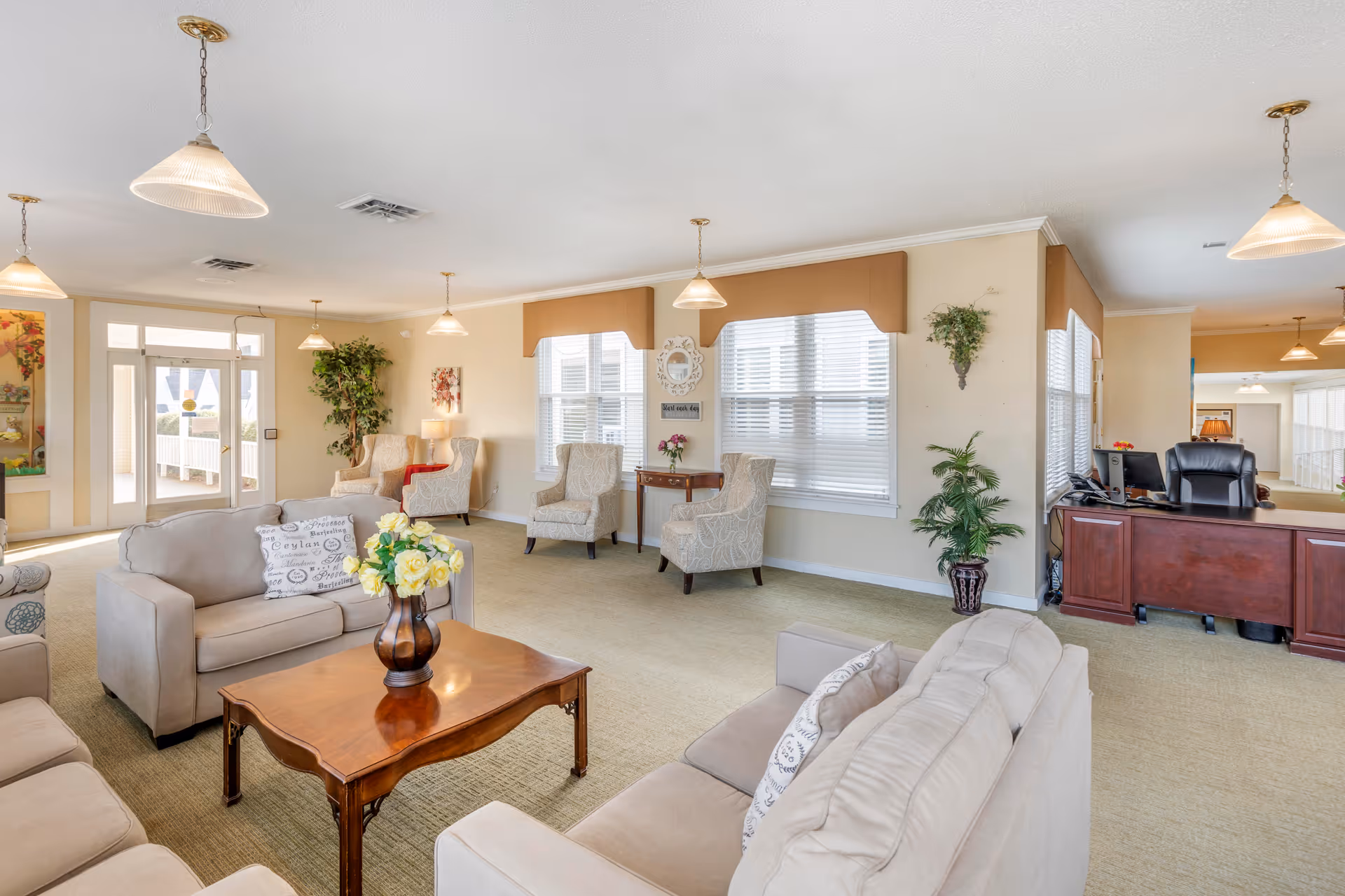 A bright and spacious senior living facility common area with beige sofas and armchairs arranged around a wooden coffee table with a vase of yellow flowers. The room has large windows with blinds, several hanging pendant lights, and decorative plants. A wooden reception desk with office equipment is visible on the right side.