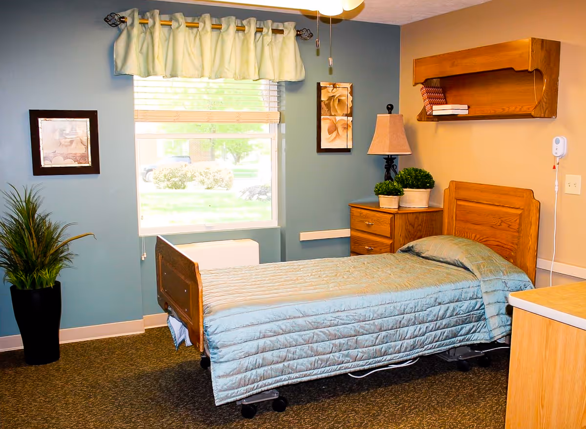 A tidy patient bedroom with a single hospital-style wooden bed, nightstand and lamp, window with a valance, and potted plants.