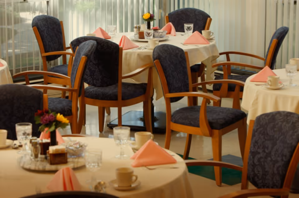 A dining room with round tables covered in beige tablecloths, each set with pink folded napkins, cups, glasses, and small flower arrangements. The chairs have wooden frames and dark patterned upholstery. Vertical blinds cover the windows in the background.