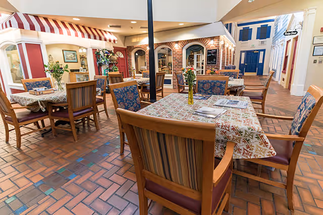 Interior view of a senior living facility common area with multiple wooden tables and chairs. The tables are covered with patterned tablecloths and decorated with small flower vases. The space features a brick floor and walls with storefront-style decor, including awnings and window displays, creating a cozy and inviting atmosphere.