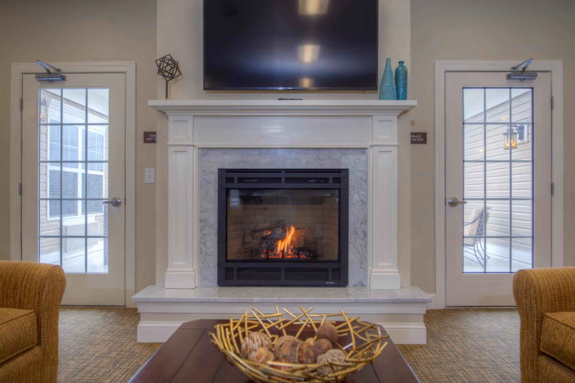 A cozy living room area featuring a lit fireplace with a marble surround and white mantel. Above the fireplace is a mounted flat-screen TV. On either side of the fireplace are glass-paneled doors leading outside. Two brown upholstered chairs are partially visible on the left and right edges of the image. A wooden coffee table with a decorative bowl filled with natural elements is in the foreground.